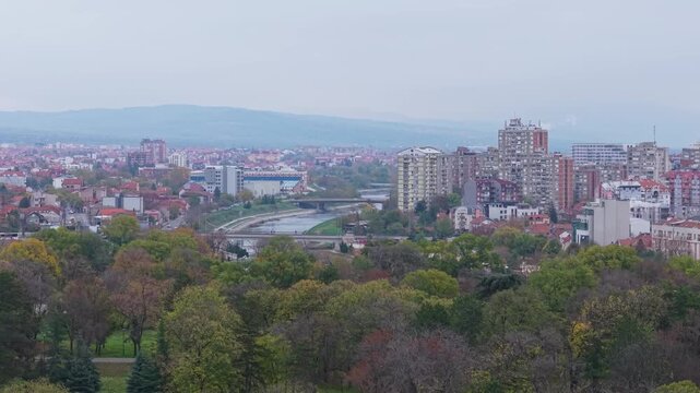 Drone ascends from behind trees, gradually revealing the sprawling cityscape of Ni&scaron;, Serbia, blending greenery with urban rooftops and streets in a cinematic aerial view.