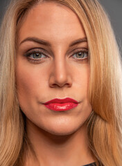 A close-up studio portrait of a blonde woman with blue eyes wearing bold red lipstick and professional makeup against a neutral grey background.