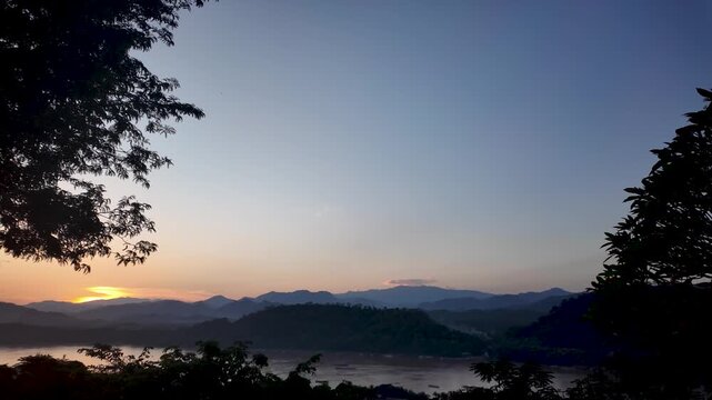 Scenic view of Luang Prabang sunset over the Mekong River from Mount Phousi in Laos with silhouette trees and mountain range in the background.