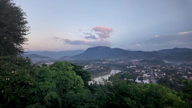 Scenic view of Luang Prabang cityscape overlooking the Mekong River from Mount Phousi at dusk featuring lush greenery mountains and a twilight sky in Laos Southeast Asia.
