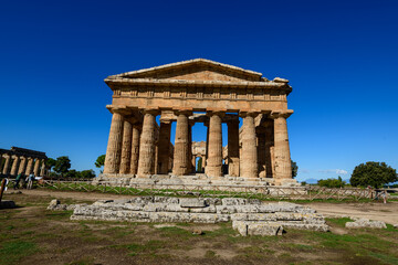 Obraz premium The well-preserved Doric columns of the Temple of Neptune rise against a vivid blue sky at the archaeological site of Paestum, Campania, Italy. Sunlight highlights the weathered stone and classical