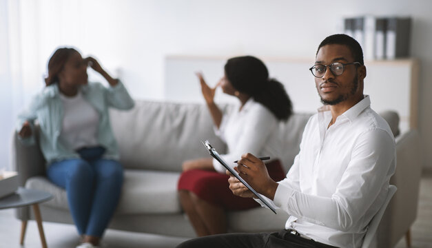 Stressed black male psychologist and angry LGBT lesbian couple fighting during session at office, copy space. Homosexual spouses having conflict on consultation with psychotherapist, banner