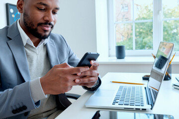 Young adult Black man sitting at desk using smartphone while working on laptop in modern office setting, focusing on device screen with natural light coming through window