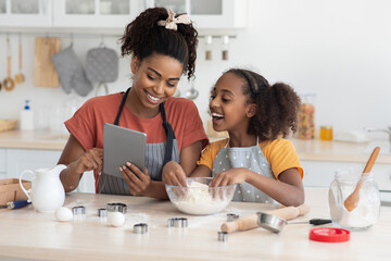 Happy black mummy and daughter cooking at home together, wearing aprons, standing by kitchen table...