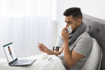 A sick Middle Eastern man sits in bed wearing a scarf and holding tissue while using a laptop for a...