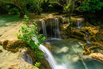 Bright sunlight illuminates a tiered limestone cascade flowing into a clear turquoise pool at Kuang Si Falls in northern Laos. Lush green foliage and mossy rocks surround the tranquil water in this