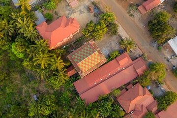 Overhead perspective of red-tiled buildings and a checkered rooftop surrounded by lush tropical trees in Krong Preah Vihear, northern Cambodia. Warm sunlight highlights the mix of greenery and urban