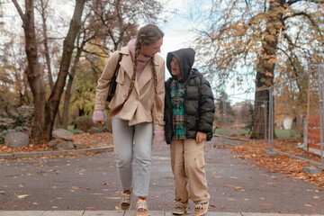 Obraz premium Woman walks with boy during autumn day at outdoor park pathway. Dry leaves cover ground while trees and fencing line scene, showing warm companionship with layered clothing and calm movement.