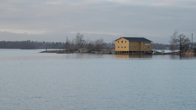The yellow wooden clubhouse of Melaveikot ry canoeing club located on the rocky Ourit island near Hietaranta beach in Helsinki, Finland, under overcast autumn sky.