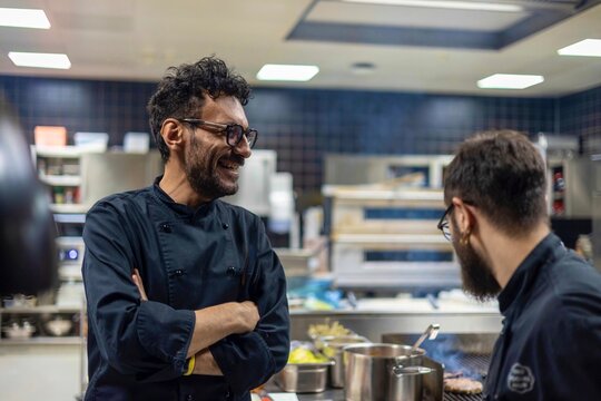 Two chefs in black uniforms smiling and talking in a professional kitchen setting - Powered by Adobe