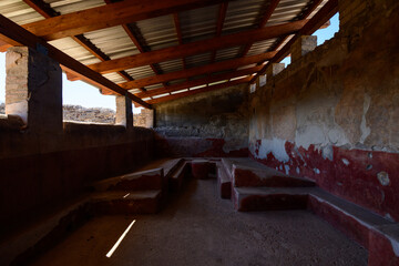 Fototapeta premium Stone benches line an ancient Roman dining room with faded red frescoes and weathered plaster in the House of the Lararium of Achilles, Pompeii, Italy. Diffused daylight enters through open windows