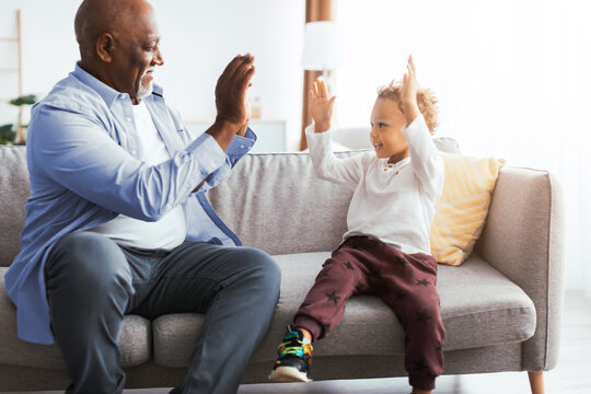 A man and a child sit on a couch in a well-lit living room. They are playing a hand-clapping game, smiling and enjoying their time together. Their interaction shows joy and connection. - Powered by Adobe