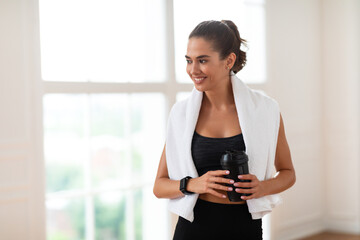 Portrait of sporty beautiful smiling woman in sportswear and white towel on neck holding shaker...