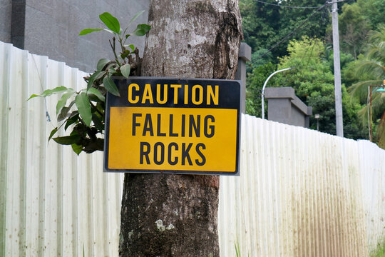 ellow and black Caution Falling Rocks sign mounted on a tree trunk next to a white corrugated fence in a lush green area.