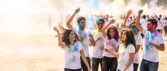Group of happy young people celebrating Holi festival with colorful powder. Friends dancing and laughing outdoors during Indian spring festival. Wide banner with copy space