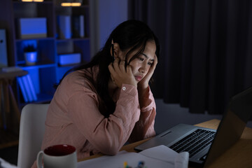 Stressed young Asian woman working late night on laptop feeling exhausted and overwhelmed with deadline burnout at home office desk