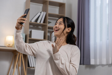 Happy and cheerful young asian woman with smile holding smartphone while making video call online with friend in living room at home, female having fun taking selfie photo with mobile phone