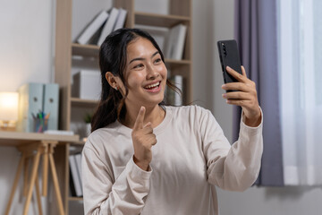 Happy young asian woman holding smartphone video call and pointing finger with cheerful smile expression in home office living room while communicating online
