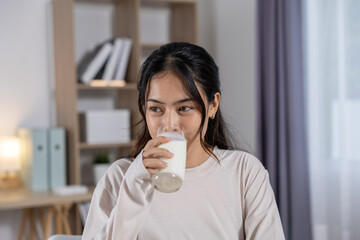 Calm young beautiful asian woman holding glass milk drinking fresh beverage with happy smile face feeling healthy energetic at home living room morning relax