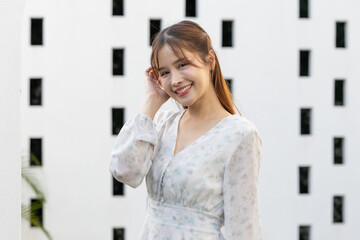 Cheerful young asian woman standing outdoor smiling brightly wearing white floral dress touching hair feeling happy confident relax against modern textured architecture wall