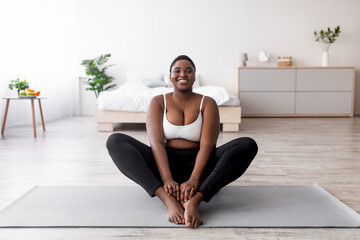 Obraz premium Cheerful plus size black woman doing yoga on sports mat at home, free space. Happy overweight Afro lady working out indoors, losing weight, leading healthy lifestyle indoors