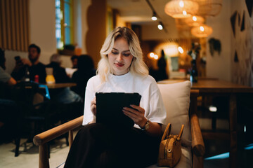 Young blonde freelancer sitting in café working on tablet with stylus, focusing on creative digital design, remote collaboration and online productivity in cozy workspace with modern lighting