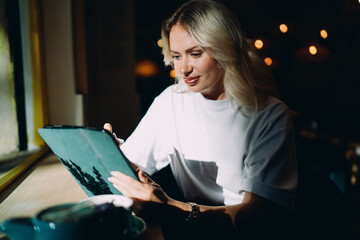 Focused young woman using digital tablet with stylus near window in soft sunlight, symbolizing modern remote workflow, digital design, and creative freelance productivity in connected workspace