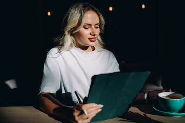 Woman using digital tablet with stylus while drinking tea in modern café, representing concept of connected freelance lifestyle, flexible remote work, and professional digital creativity