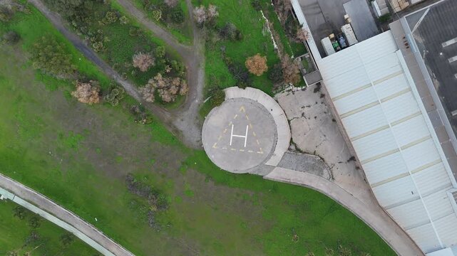 Top-down aerial view of a helicopter landing pad near an industrial building, drone perspective showing helipad markings, green landscape and access roads