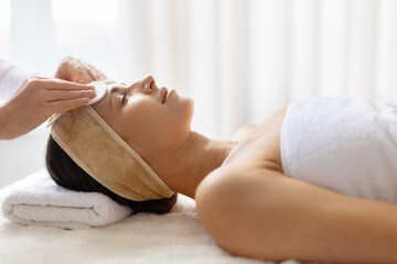 Woman enjoying facial cleansing treatment at spa, eyes closed and relaxed, minimal interior...