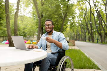 Happy handicapped black man in wheelchair using laptop for online job at cafe in city park, having business meeting on web, outdoors. African American disabled freelancer guy working on remote project