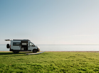 White Camper Van Parked On Green Grass By The Beach With Surfboard In Open Daylight