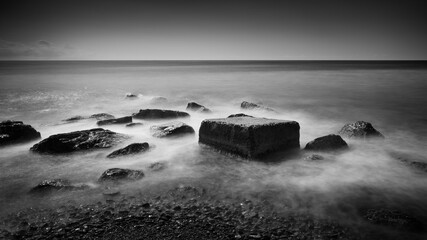 Black and white image of a rocky shore with a large rock in the middle.  water is calm, image dramatic, long exposure, smooth white water, black rocks