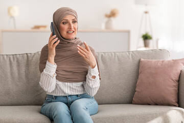 Obraz premium Happy Mature Middle-Eastern Female Having Phone Conversation Talking By Cellphone Sitting On Couch At Home, Wearing Hijab. Arab Housewife Chatting Using Smartphone Indoor. People And Gadgets