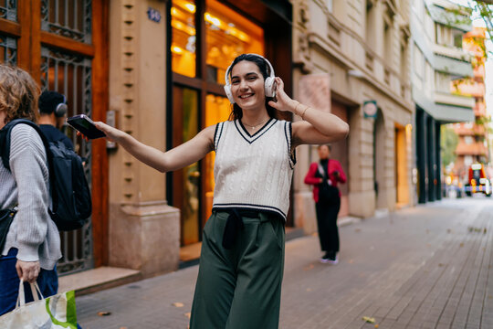 Woman smiling and dancing with headphones in the street while holding smartphone — celebrating joyful tech interaction through sound, movement, and self-expression. - Powered by Adobe