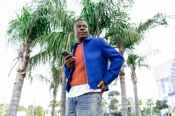 A young African man stands outdoors near palm trees, holding a smartphone. He wears a blue jacket over an orange sweater and jeans.