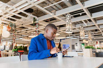 A young Black man using a phone and holding a cup and sits at a table in a modern cafe, . The interior features contemporary decor and bright lighting.