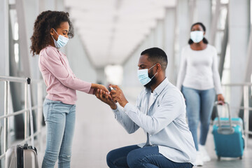 Fototapeta premium Health Care And New Normal Protection Concept. Black Father Spraying Sanitizer On Daughter's Hands Cleaning Arms In The Airport Terminal Indoors, Wearing Protective Medical Masks For Covid Prevention