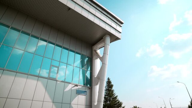 Glass wall rises near white support column under deep roof overhang. Steel brace anchors corner while window grid reflects blue sky