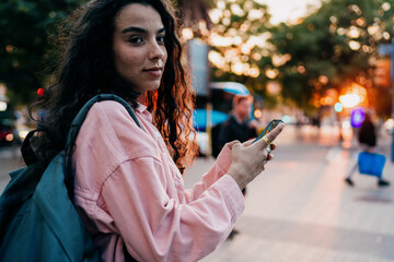 Young woman looks at camera while holding phone, expressing digital self-assurance, mobile strength and contemporary identity shaped by constant online connection and urban rhythm.