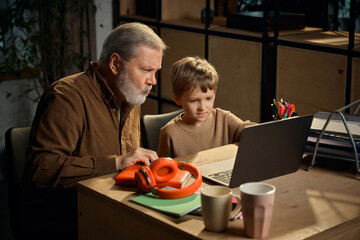 Young boy mentoring senior man using laptop at home desk. Concept of reverse mentorship, digital skills learning, technology adoption, family knowledge exchange, modern education.