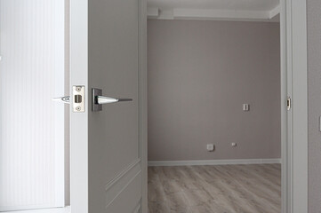 partially open white interior door with a sleek metallic handle and lock mechanism. Behind it — a minimalist room with light grey walls, light wood-patterned flooring, and a few wall sockets switches