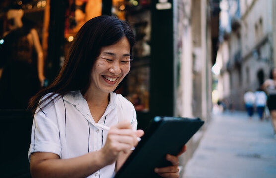 Asian woman smiling while using digital tablet with stylus, showing delight in creative process, digital autonomy and confident freelance mindset in mobile work.