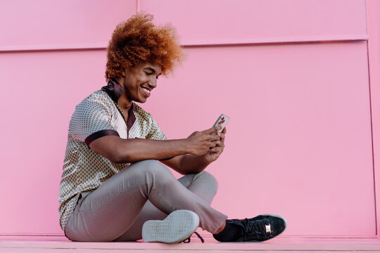 Young African American man with curly red hair sitting casually on ground, smiling while looking at smartphone screen, portraying relaxed tech use and joyful online engagement outdoors. - Powered by Adobe