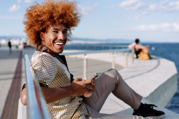 Joyful African American man with voluminous ginger afro reclining on promenade ledge, checking smartphone, wearing silky patterned shirt and plaid trousers, representing connected coastal lifestyle.
