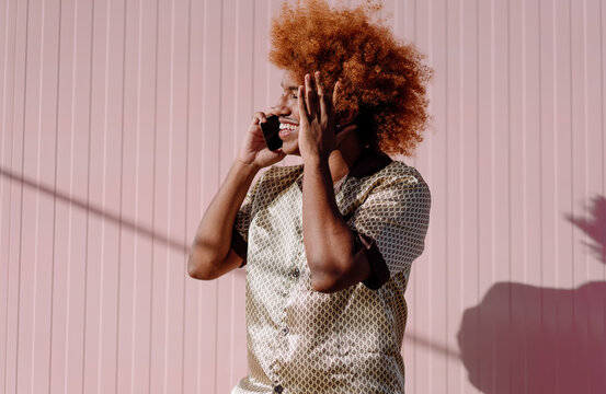 Smiling African American man with curly red afro holding smartphone, hands near head, wearing patterned silk shirt, representing excitement and engagement in digital communication.