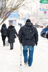 Senior man in black puffer coat and beanie walking slowly through falling snow with cane and...