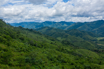 Fototapeta premium Lush green hills and densely wooded mountain ridges extend into the distance beneath a sky filled with textured clouds in the countryside between Phonsavan and Phou Khoun, Laos. The landscape