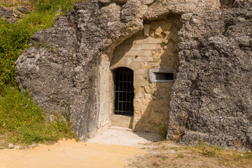 Close-up of a weathered stone doorway with metal bars set into a rugged rock face at Fort de Douaumont. The historic entrance is surrounded by rough textures, sparse grass, and sunlit earth in the