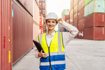 Smiling female logistics manager in a hard hat and safety vest holding a tablet, standing in a shipping container terminal. Professional port inspector or engineer overseeing global freight.
